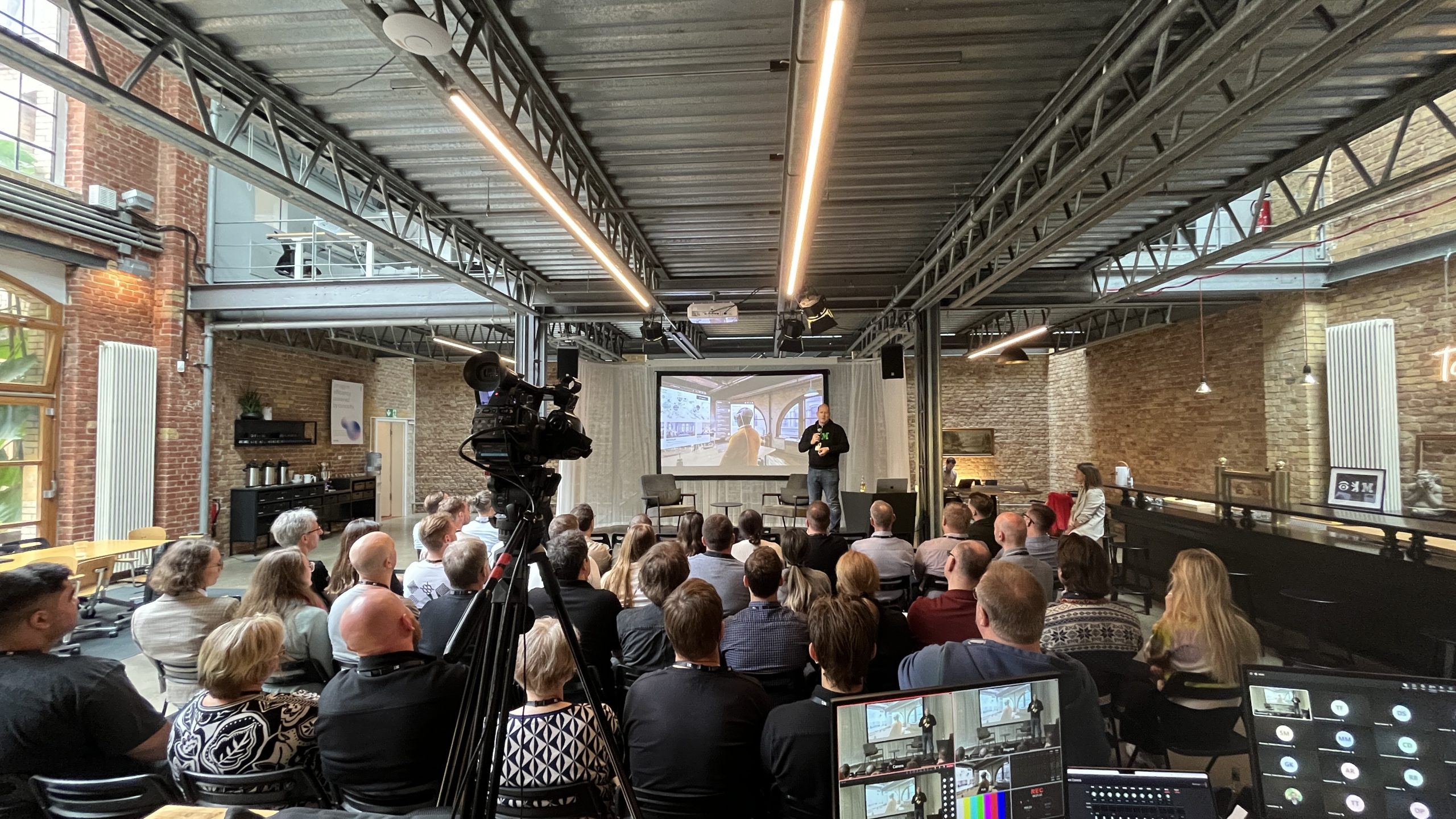 Viele Menschen sitzen auf Stühlen in einer Halle. Vorne ist eine Kamera und ein Monitor zu sehen und im Hintergrund eine Bühne mit einer großen Leinwand.