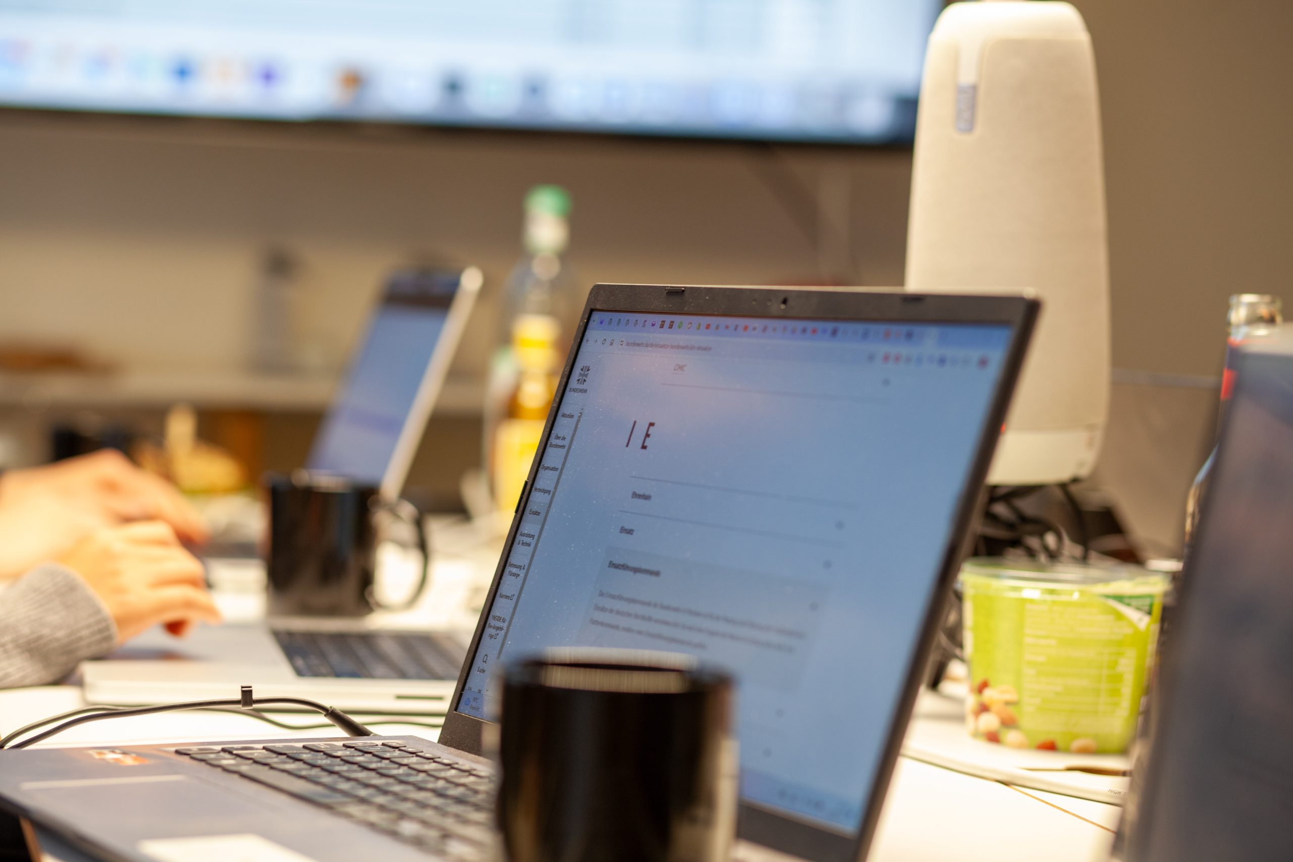 Auf einem Konferenztisch stehen viele Laptops, Kaffeetassen und ein Konferenzmikrofon. Im Hintergrund ist eine Präsentation an die Wand projiziert.