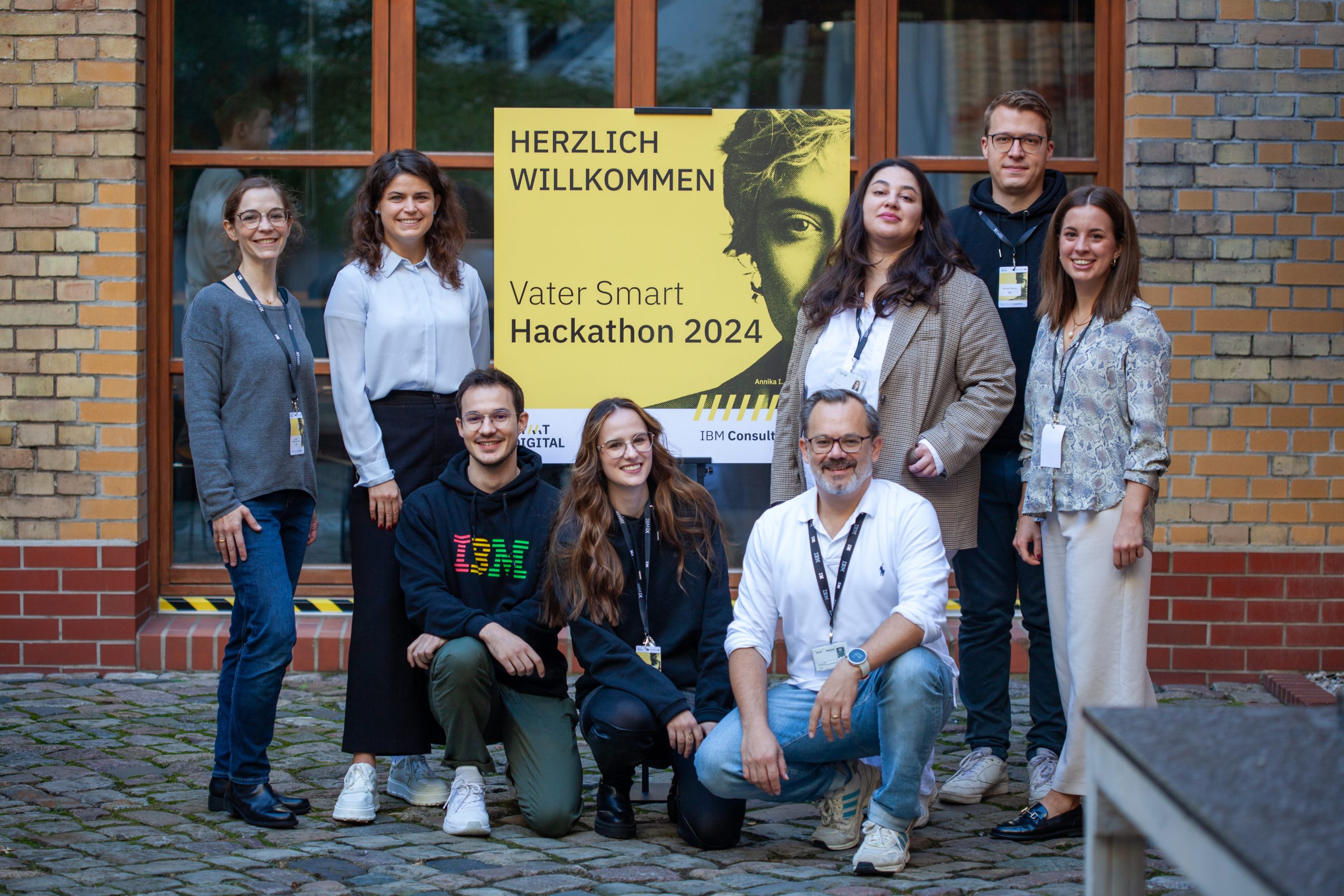 Acht Menschen stehen und hocken rund um ein Schild vor einem Backsteingebäude. Das Schild trägt die Aufschrift: Herzlich willkommen, Vater Smart Hackathon 2024.